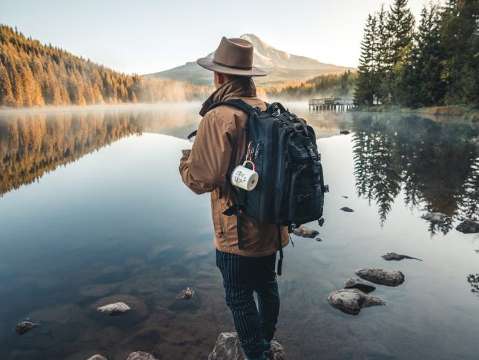 man in brown jacket and brown hat standing on rock near lake